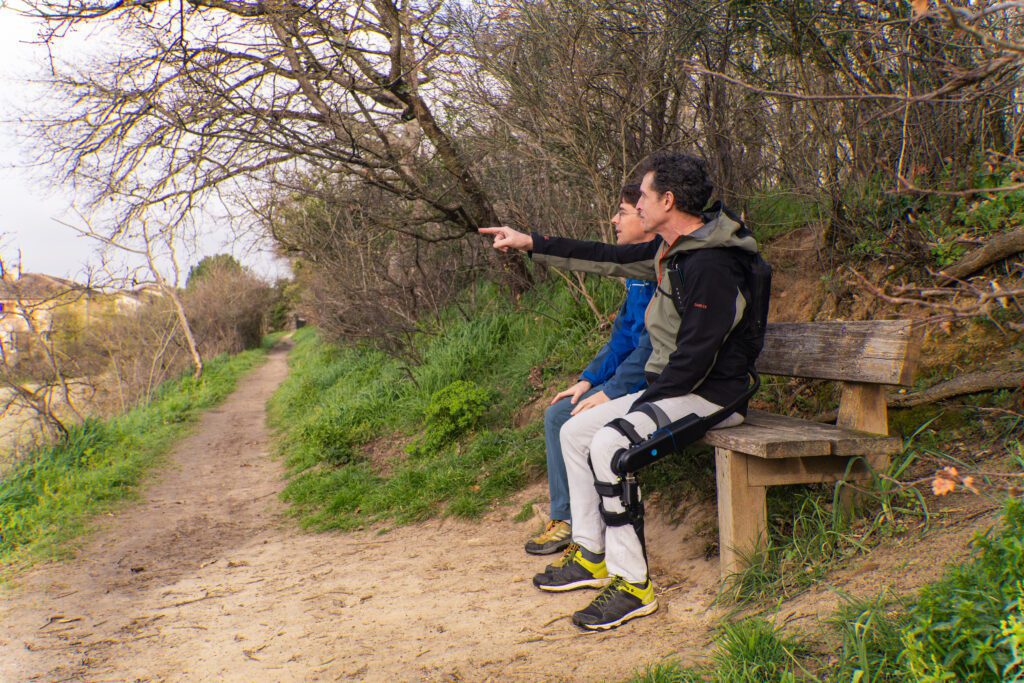 Two men sitting on an outdoor bench along a trail. The man on the left is wearing the DREEVEN robotic knee brace.