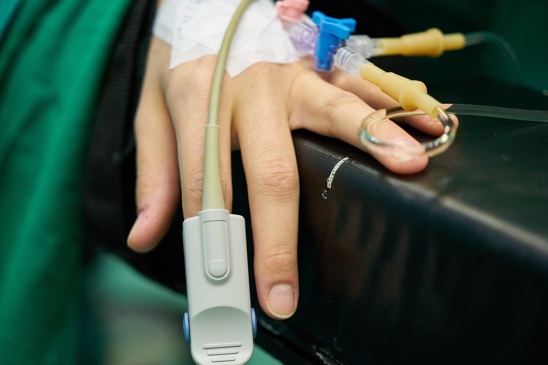 Upclose image of a hand resting on a dark surface. The back of the hand has an IV in, and the index finger has a pulse oximeter on it.